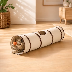 Peut inclure: Un tunnel pour chat cylindrique en tissu blanc cassé avec des bordures marron, doté d'une ouverture circulaire sur le dessus. Un chat tigré est à l'intérieur du tunnel et tend une patte pour jouer avec une petite balle orange suspendue.