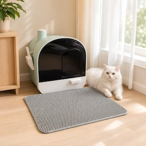 May include: A sage green and black enclosed cat litter box with a white ribbed base, a matching scoop attached to the side, and a rectangular gray textured litter mat placed in front, accompanied by a fluffy white cat sitting beside it.