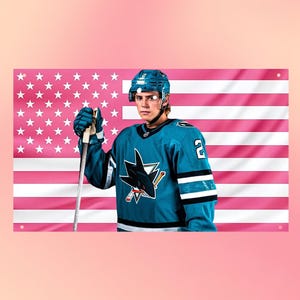 May include: A hockey player wearing a teal jersey with a shark logo and the number 2, a teal helmet, and gloves, holding a hockey stick. The background is a flag featuring horizontal pink and white stripes with a field of white stars.