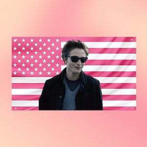 Puede incluir: Una bandera de pared rectangular con un patrón de bandera estadounidense en rosa y blanco, con una impresión fotográfica central de una persona de cabello oscuro que usa gafas de sol negras, una camiseta gris y una chaqueta negra.