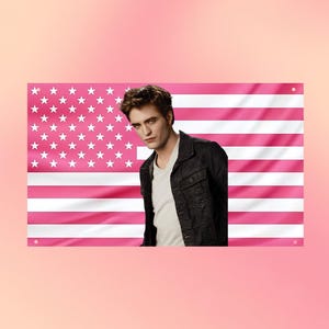 Puede incluir: Un estandarte de pared rectangular con un patrón de bandera estadounidense en rosa y blanco, que muestra el retrato de una persona de cabello oscuro con una camiseta blanca y una chaqueta negra. Tiene cuatro ojales metálicos en las esquinas.