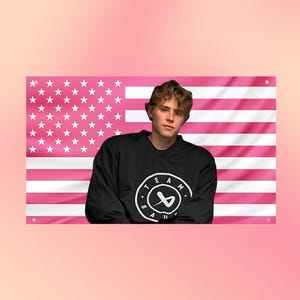 Könnte beinhalten: Ein schwarzes Langarm-Sweatshirt mit einem kreisförmigen weißen Logo, das den Text TEAM BAE und eine stilisierte Grafik in der Mitte zeigt. Das Sweatshirt ist vor einer rechteckigen Flagge mit einem rosa-weißen US-Flaggenmuster platziert.