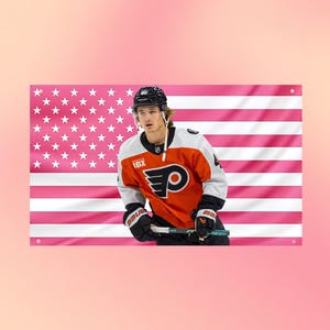 Puede incluir: Un estandarte de pared rectangular que muestra a un jugador de hockey con una camiseta naranja, negra y blanca con el logotipo de los Philadelphia Flyers y el texto IBX, sosteniendo un palo de hockey, sobre un fondo de bandera estadounidense rosa y blanca.