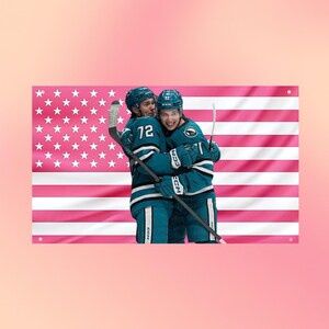 Könnte beinhalten: Eine rechteckige Wandflagge mit einem rosa-weißen amerikanischen Flaggenmuster im Querformat. Zwei Eishockeyspieler in türkisfarbenen und weißen Trikots mit der Nummer 72 umarmen sich und halten einen Hockeyschläger vor der Flagge.