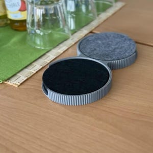 Untersetzergefäß in Grau mit schwarzen Filzuntersetzern auf einem Holztisch, daneben Flasche und Streuer
