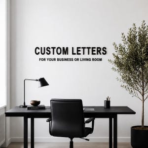 Puede incluir: Letras negras montadas en la pared que deletrean CUSTOM LETTERS FOR YOUR BUSINESS OR LIVING ROOM en una fuente sans-serif. Las letras est&aacute;n fijadas en una pared blanca lisa sobre un escritorio negro, una silla de oficina, una l&aacute;mpara y una planta.