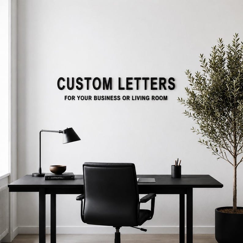 Puede incluir: Letras negras montadas en la pared que deletrean CUSTOM LETTERS FOR YOUR BUSINESS OR LIVING ROOM en una fuente sans-serif. Las letras est&aacute;n fijadas en una pared blanca lisa sobre un escritorio negro, una silla de oficina, una l&aacute;mpara y una planta.
