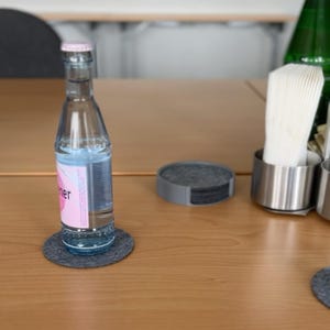 Runder Untersetzerhalter in Grau mit Stone-Grey-Filzuntersetzern auf einem Holztisch, eine Wasserflasche steht auf einem der Untersetzer