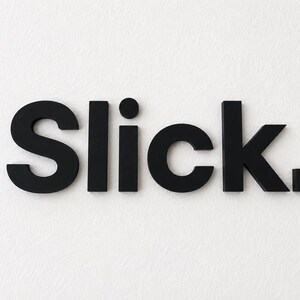 Puede incluir: La palabra Slick. representada en letras negras, negritas y tridimensionales. Los caracteres est&aacute;n dispuestos horizontalmente, con un punto distintivo sobre la letra i y un punto cuadrado al final de la palabra.