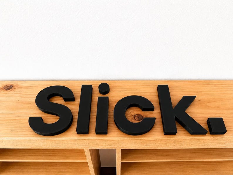 Puede incluir: Un conjunto de letras tridimensionales en negro mate que forman la palabra "Slick." con un punto al final. Las letras est&aacute;n dispuestas en una l&iacute;nea horizontal y presentan una tipograf&iacute;a sans-serif limpia con un acabado liso y s&oacute;lido.