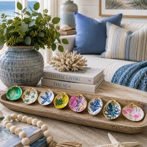May include: A long, narrow, natural wood dough bowl containing eight individual oyster shells, each hand-painted with distinct coastal motifs including palm trees, a blue starfish, pink flamingos, an octopus, and vibrant tropical foliage.