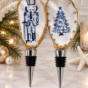 Könnte beinhalten: Zwei Flaschenverschlüsse mit Austernschalen mit goldfarbenen Rändern. Eine Schale zeigt einen blau bemalten Nussknacker mit silbernen Perlenakzenten, die andere einen blau bemalten Weihnachtsbaum, verziert mit kleinen weißen und silbernen Perlen.