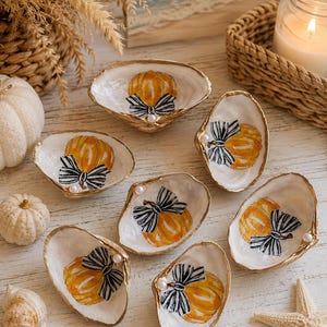 May include: A collection of six decorative oyster shells, each featuring a hand-painted orange pumpkin with a black and white striped bow. The edges of the shells are trimmed in gold, and each shell is accented with a small white pearl bead.