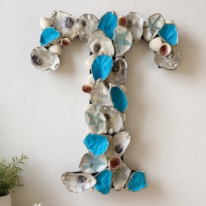 May include: A decorative letter T covered in a variety of natural seashells and starfish. The arrangement features white, tan, and brown oyster shells interspersed with vibrant turquoise painted shells and light blue starfish accents throughout.