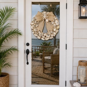 May include: A circular wreath constructed from numerous natural oyster shells, featuring a large tan burlap bow at the top center. A small white starfish and a single small seashell are attached to the center of the bow as decorative accents.