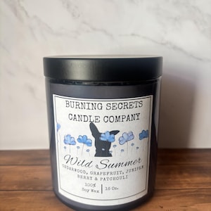 May include: A 16 oz. black jar candle with a black lid and a white label. The label reads Burning Secrets Candle Company, Wild Summer, Cedarwood, Grapefruit, Juniper, Berry & Patchouli, and 100% Soy Wax. It features a black dog silhouette and blue flowers.