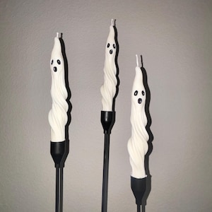 Peut inclure: Trois bougies effilées blanches en forme de fantômes avec des corps torsadés et des yeux et bouches peints en noir. Chaque bougie est placée dans un bougeoir en métal noir fin. Les bougies ont une texture de cire lisse et une mèche blanche.