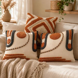 May include: Three square throw pillows with cream-colored fabric bases. Two pillows feature dark gray and burnt orange tufted geometric shapes and braided trim. One pillow displays a large, tufted burnt orange and cream diamond pattern.