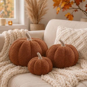 May include: Three round, rust-colored pumpkin-shaped pillows made of textured, fuzzy fabric. Each pillow features a short, light-colored stem at the top. The pillows vary in size and are arranged together, showcasing a soft, plush, and rounded appearance.