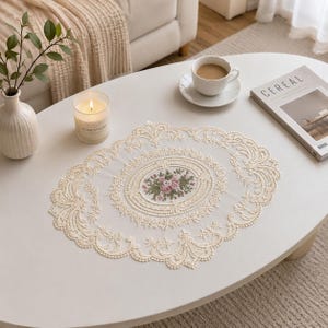 May include: An oval-shaped cream-colored lace doily featuring an intricate embroidered floral center with pink roses and green leaves, surrounded by a detailed scalloped border with elaborate openwork patterns and decorative stitching.