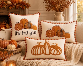 May include: Three cream-colored throw pillows with orange pom-pom trim. Two pillows feature embroidered pumpkins and the text "It's Fall y'all" on the square one. The third rectangular pillow displays three embroidered orange pumpkins.
