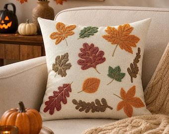 May include: A square throw pillow with a cream-colored fabric base featuring an embroidered pattern of various autumn leaves in shades of orange, red, brown, and green. The textured leaf designs are scattered across the front surface.