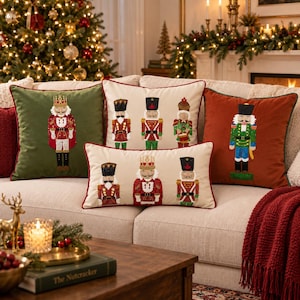 May include: A collection of four decorative throw pillows featuring embroidered nutcracker figures. One pillow is olive green, one is rust orange, and two are cream-colored with red piping. The designs include various nutcracker soldiers.