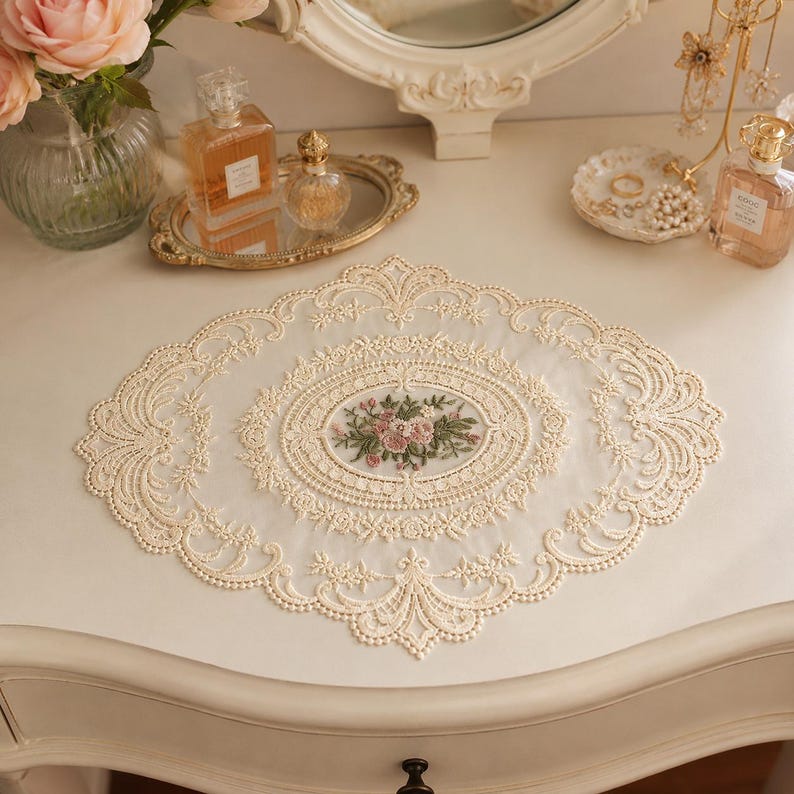 May include: An oval-shaped cream-colored lace doily featuring intricate embroidery of pink and green flowers in the center. The doily has a delicate scalloped edge with detailed floral patterns and a sheer mesh fabric base, creating a decorative mat.