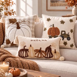 Puede incluir: Tres cojines decorativos con motivos otoñales. Un cojín cuadrado tiene una calabaza blanca sobre un fondo de cuadros beige. Otro cojín cuadrado muestra un tractor con una calabaza. Un cojín rectangular muestra calabazas y la palabra autumn.