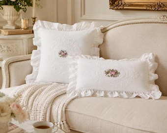 May include: Two white throw pillows with ruffled edges and central floral embroidery. The square pillow features a circular lace-like pattern around a small rose bouquet, while the rectangular pillow has an oval lace-like border around roses.