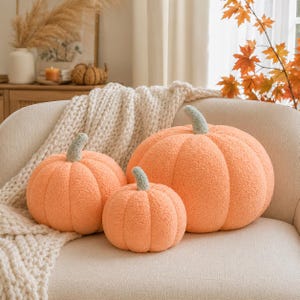 May include: Three plush pumpkin-shaped pillows in varying sizes, crafted from a soft, textured orange fabric. Each pumpkin features a distinct, rounded ribbed design and a short, sage green stem attached to the top center of the rounded body.