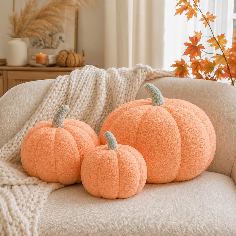May include: Three plush pumpkin-shaped pillows in varying sizes, crafted from a soft, textured orange fabric. Each pumpkin features a distinct, rounded ribbed design and a short, sage green stem attached to the top center of the rounded body.
