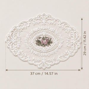 May include: An oval white lace doily featuring intricate floral embroidery in the center with pink and green thread. The lace border has a detailed scalloped edge. Dimensions are 14.57 inches in width and 11.42 inches in height as indicated.