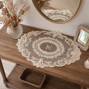 May include: An oval-shaped cream-colored lace doily featuring intricate floral embroidery in the center with pink and green thread. The doily has a scalloped edge with detailed openwork patterns and is crafted from a sheer, lightweight fabric.