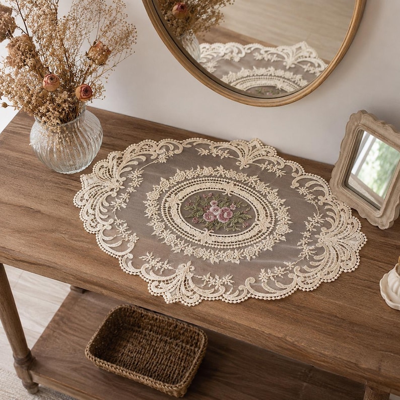 May include: An oval-shaped cream-colored lace doily featuring intricate floral embroidery in the center with pink and green thread. The doily has a scalloped edge with detailed openwork patterns and is crafted from a sheer, lightweight fabric.