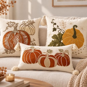 Peut inclure: Trois coussins décoratifs couleur crème ornés de broderies de citrouilles dans des tons orange, jaune et vert. Les coussins carrés ont des pompons aux coins, tandis que le coussin rectangulaire présente trois citrouilles et des pompons.