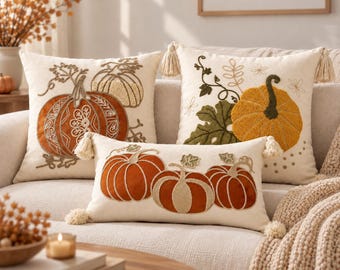 May include: Three cream-colored throw pillows featuring embroidered pumpkin designs in shades of orange, yellow, and green. The square pillows have corner tassels, while the rectangular pillow features three pumpkins and tassels on each side.
