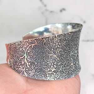 May include: A wide silver cuff bracelet featuring an intricate, embossed floral pattern across the entire outer surface. The metal has a polished finish on the interior and a darkened, oxidized texture within the recessed areas of the floral design.