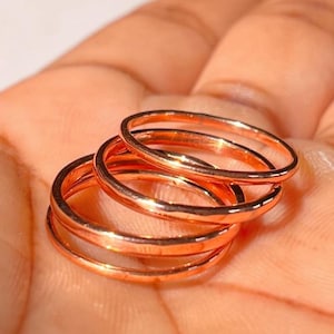 May include: A set of three thin, polished rose gold-coloured metal rings with a smooth, rounded surface. The rings are stacked together, showcasing a metallic sheen and a simple, circular band design that is uniform in thickness and colour throughout.