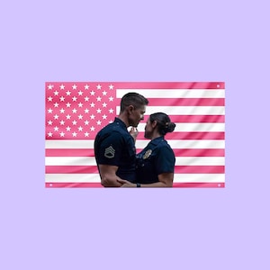 May include: A rectangular wall flag featuring a pink and white American flag design. In the center, a police officer in a dark blue uniform stands face-to-face with another officer in a dark blue uniform, both looking at each other.