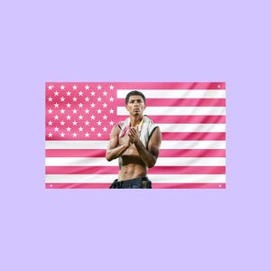 Può includere: Uno stendardo da parete rettangolare con un design della bandiera americana in rosa e bianco. Al centro della bandiera c'è una stampa fotografica di un atleta a torso nudo con capelli scuri corti, che indossa un asciugamano bianco al collo.