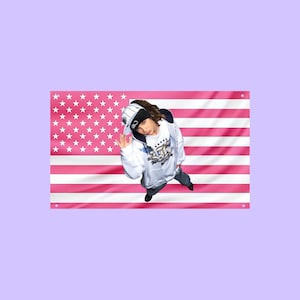 Puede incluir: Una bandera de pared rectangular con un patrón de bandera estadounidense en rosa y blanco. Centrado en la bandera hay un gráfico de una persona con una sudadera blanca con cremallera y detalles azules, una gorra de béisbol negra y blanca y pantalones oscuros.