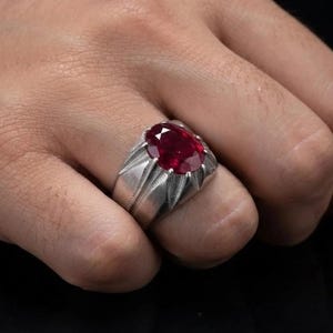 Puede incluir: Un anillo de plata con una gran piedra preciosa de color rojo intenso y corte ovalado, montada en una base de garras. La banda tiene un acabado de metal cepillado con detalles de ranuras verticales que bajan desde el engaste.