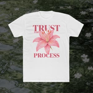 Op de afbeelding: Een wit t-shirt met korte mouwen en een roze leliebloemafbeelding in het midden van de borst. De tekst "TRUST THE PROCESS" is gedrukt in een roze schreeflettertype, met "TRUST" boven de bloem en "THE PROCESS" onder de bloem.