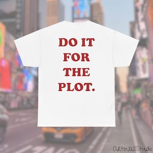 Puede incluir: Una camiseta blanca de manga corta con el texto DO IT FOR THE PLOT impreso en letras mayúsculas rojas y negritas centradas en la parte posterior. La prenda tiene un cuello redondo clásico y mangas cortas sobre un fondo urbano desenfocado.