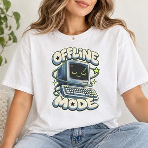 Könnte beinhalten: Ein weißes Kurzarm-T-Shirt mit einem Grafikdruck eines Vintage-Computers, dessen Bildschirm ein lächelndes Gesicht zeigt. Der Text "OFFLINE MODE" ist in einer fetten Blasen-Schriftart über und unter dem Computer abgebildet.