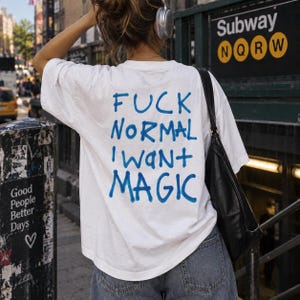 May include: A white short-sleeved t-shirt featuring the blue text "FUCK NORMAL I WANT MAGIC" printed on the back. The shirt is worn with blue denim jeans and a black shoulder bag, showcasing a casual style with a bold graphic statement on the back.