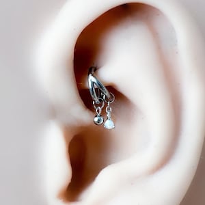 Puede incluir: Un pendiente hélix plateado con dos colgantes. Uno es una pequeña esfera y el otro es un ópalo en forma de lágrima. El pendiente está sujeto a una oreja de color claro.