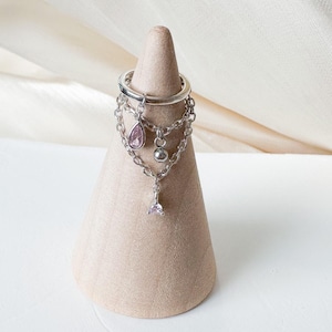 Könnte beinhalten: Ein silberfarbener Ring mit herabhängenden Ketten und rosa Edelstein-Akzenten. Der Ring wird auf einem hellbraunen, kegelförmigen Ständer vor einem weißen und cremefarbenen Hintergrund präsentiert. Der Ring hat mehrere Schichten und filigrane Details.