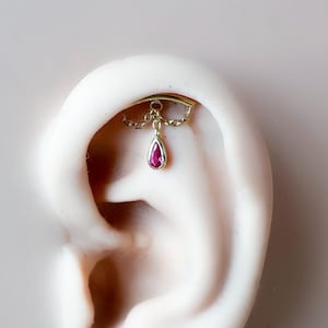 Pode incluir: Um brinco hélice dourado com uma pedra preciosa vermelha em forma de gota pendurada. O brinco é exibido em um modelo de orelha branca sobre um fundo cinza claro. A joia é delicada e elegante.